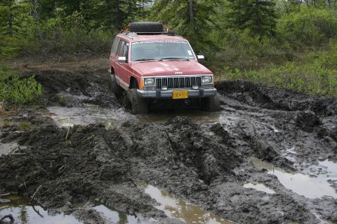 2003 Jeep TJ - A red Jeep Cherokee XJ navigates a challenging muddy trail, with its tires deep in the muck.