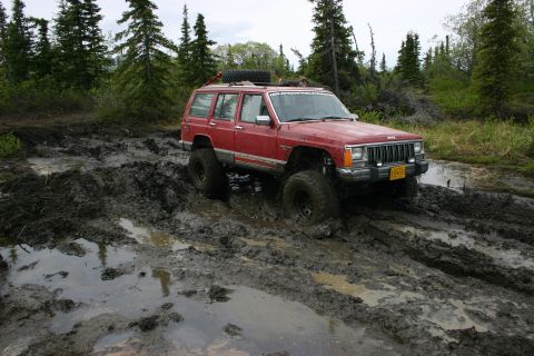 2003 Jeep TJ - A red Jeep Cherokee XJ navigates a challenging muddy terrain with its suspension lifted and off-road tires.