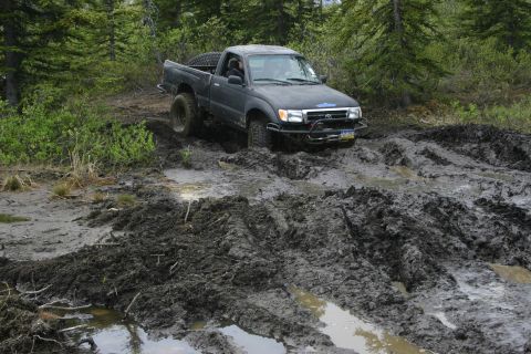 2003 Jeep TJ - Pickup truck struggling through a muddy off-road trail with trees in the background.