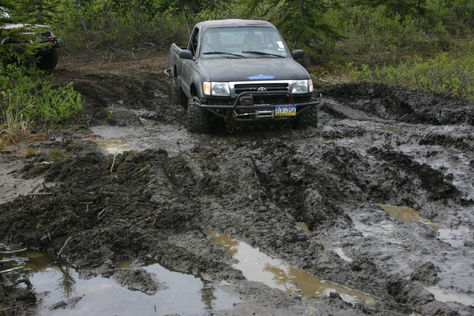 Black Toyota Tacoma struggling through deep mud on a trail. Another vehicle is visible in the background, also partially obscured by foliage