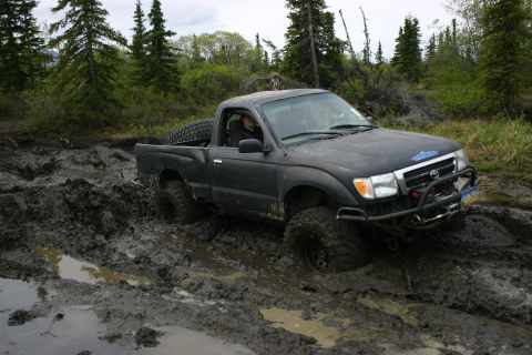 2003 Jeep TJ - A black Toyota Tacoma navigates a muddy trail, with its front grille and tires deeply embedded in the mire.