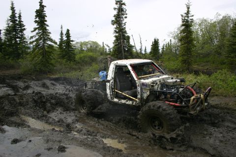 2003 Jeep TJ - A heavily modified white pickup truck navigates a treacherous muddy trail, with trees and foliage in the background.