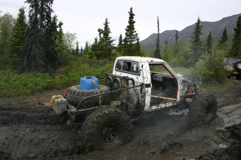 2003 Jeep TJ - An off-road pickup truck navigates a muddy trail with a spare tire and water container loaded in the back.