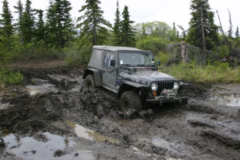 2003 Jeep TJ - A Jeep Wrangler struggles to navigate a very muddy and wet off-road trail surrounded by trees.