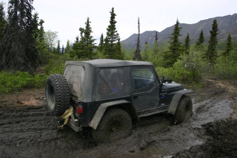 2003 Jeep TJ - A black 2003 Jeep TJ navigates a muddy trail surrounded by trees and mountains.