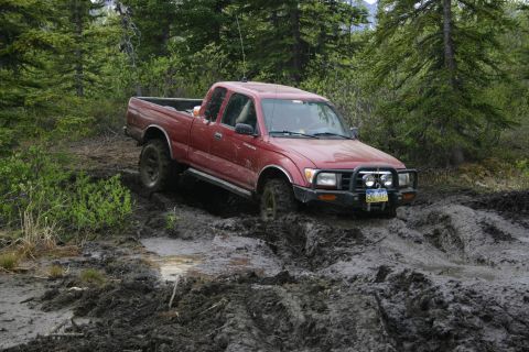 2003 Jeep TJ - This red pickup truck is navigating a challenging muddy terrain in a forest environment.