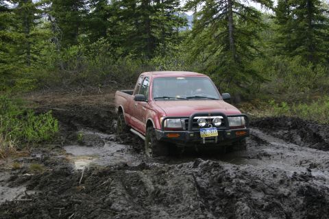 2003 Jeep TJ - A red pickup truck navigates a muddy trail surrounded by trees.