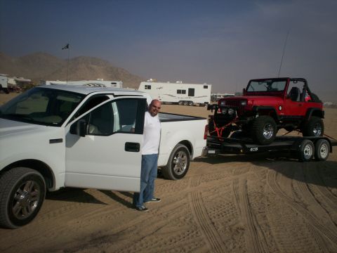 1987 Suzuki Samurai - A red Jeep Wrangler is loaded onto a trailer behind a white pickup truck in a sandy, desert environment.