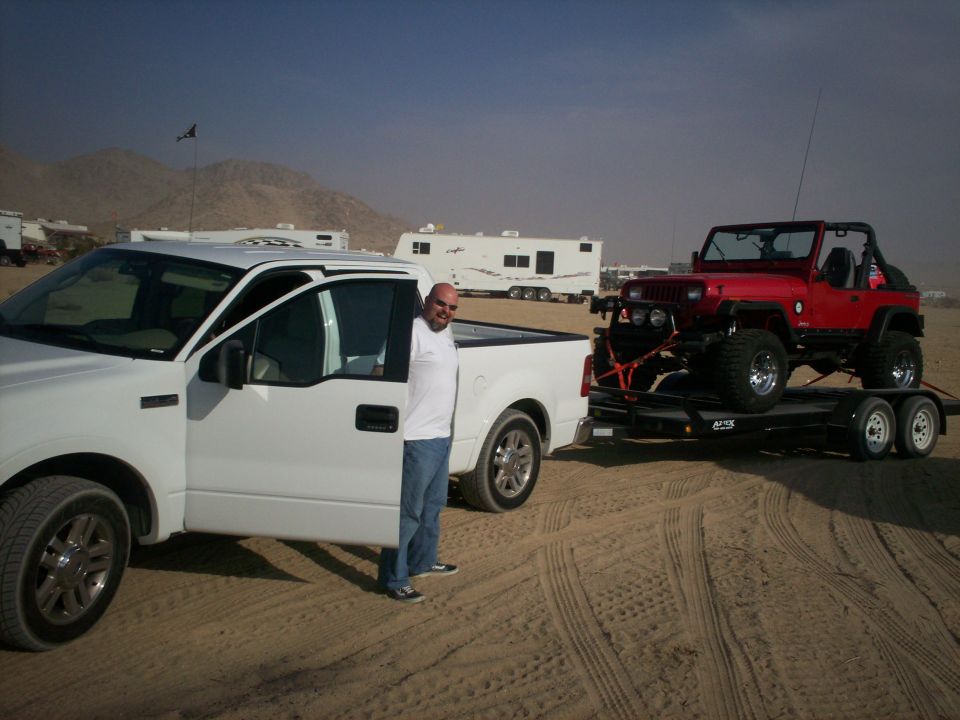 Red Jeep Wrangler on a trailer, towed by a white pickup truck, in a desert landscape