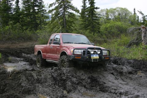 2003 Jeep TJ - A red pickup truck navigates a challenging muddy trail, its tires deep in the mire.