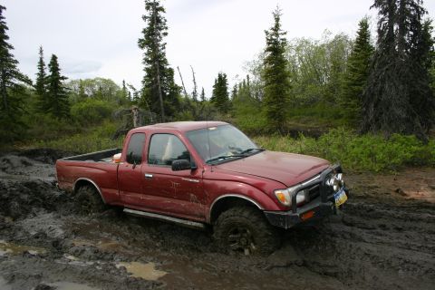 2003 Jeep TJ - A red pickup truck is stuck in thick mud on an off-road trail surrounded by trees.