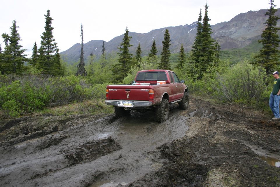 Red Toyota Tacoma pickup truck driving through mud, rear view. Mountains and trees in background