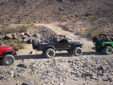 1987 Suzuki Samurai - Three off-road vehicles traverse a rocky desert path under a clear sky.