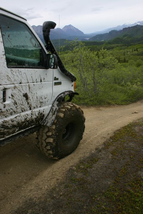 2003 Jeep TJ - A white van, heavily mud-splattered and equipped with a snorkel, navigates a dirt track through a scenic, mountainous landscape.
