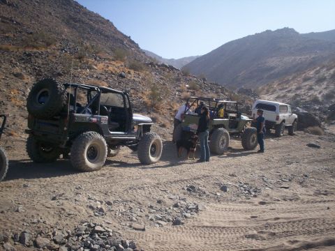 1987 Suzuki Samurai - A group of off-road vehicles are stopped on a dirt path in a desert landscape with mountains in the background.