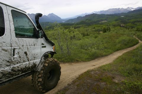 2003 Jeep TJ - A white van, covered in mud and equipped with a snorkel, navigates a dirt track through a scenic, green mountain range.