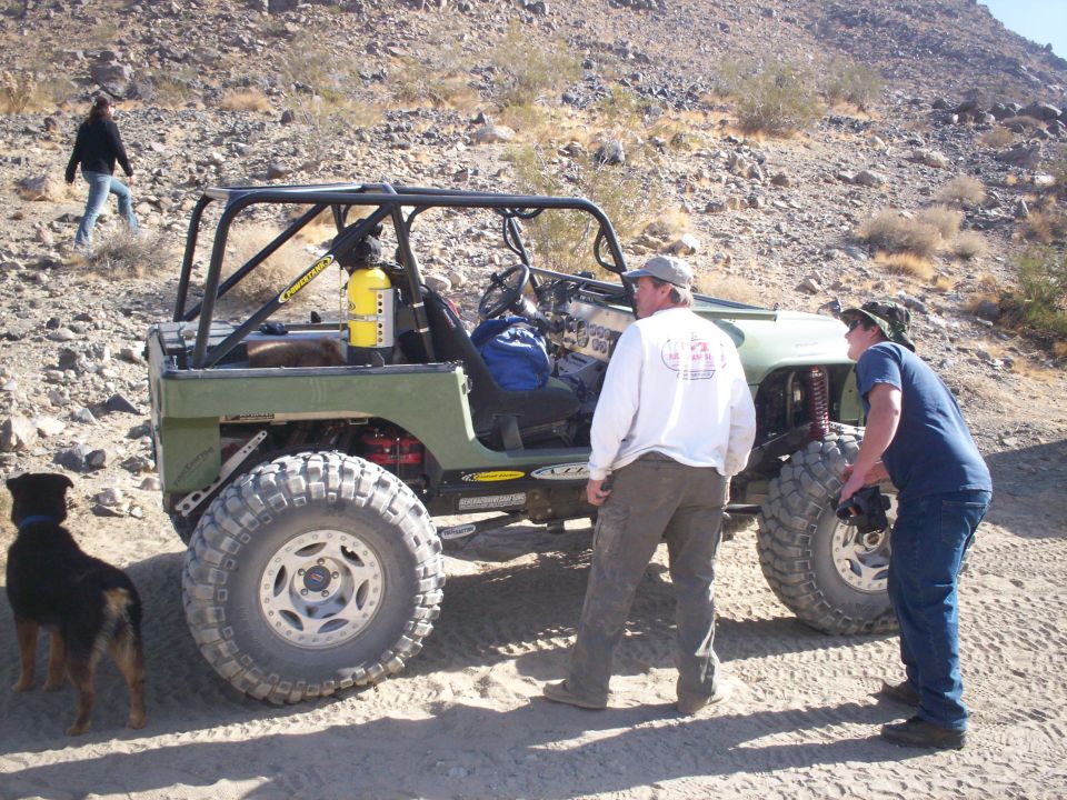 Modified Jeep with roll cage, large off-road tires, and people standing around it in a desert landscape