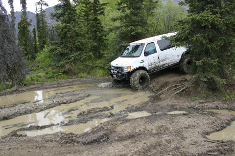 2003 Jeep TJ - A white van equipped with oversized tires tackles a challenging muddy terrain in a wooded area.