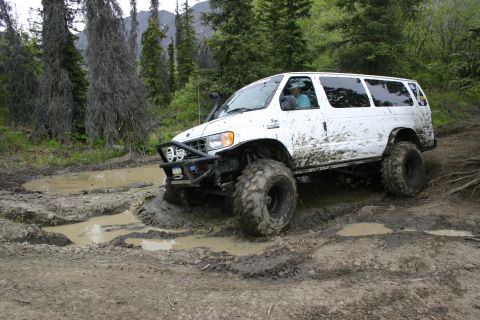 2003 Jeep TJ - A modified white van navigates a challenging muddy off-road course surrounded by trees.