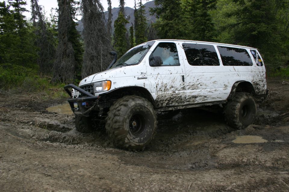 White van with large tires stuck in mud, off-road bumper, snorkel, and tree line