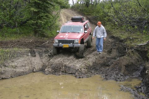 2003 Jeep TJ - A red Jeep Cherokee XJ tackles a challenging muddy off-road course, with a person walking alongside.