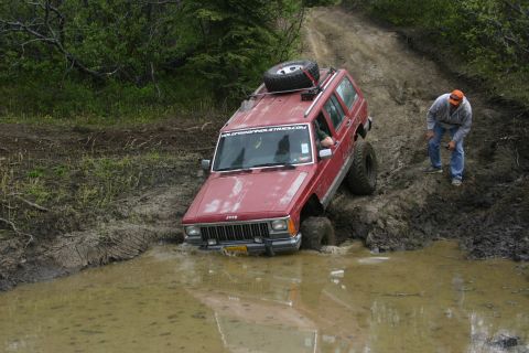 2003 Jeep TJ - A red Jeep Cherokee XJ navigates a challenging muddy trail, with a person observing the off-road excursion.