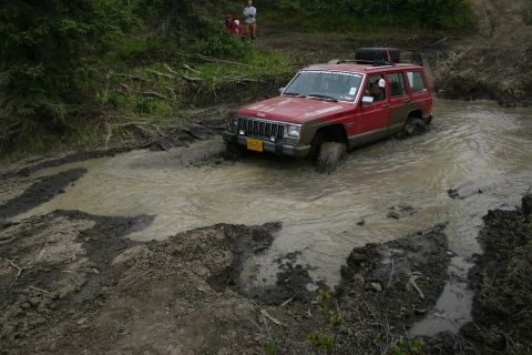 2003 Jeep TJ - A red Jeep Cherokee XJ navigates a challenging muddy terrain, with two people watching from the bank.