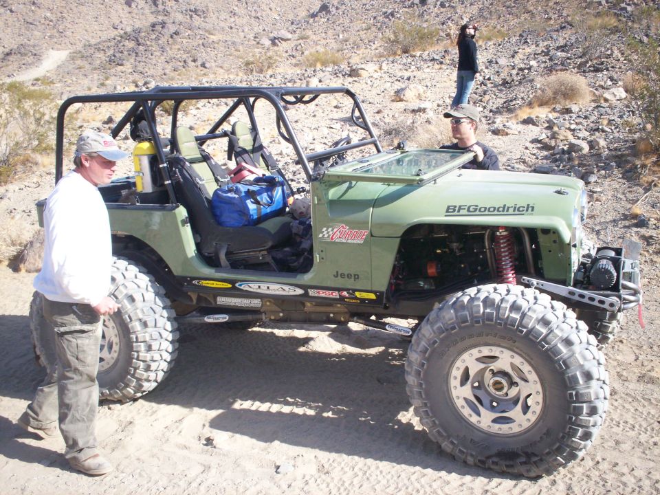 Modified green Jeep Wrangler with roll cage and large tires parked on sandy terrain. Two people stand near the vehicle, one by the door and one behind the windshield. Another person stands on a hill in the background