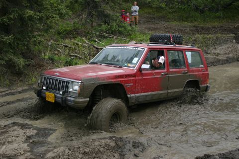 2003 Jeep TJ - A red Jeep Cherokee navigates a challenging muddy off-road course, with onlookers in the background.
