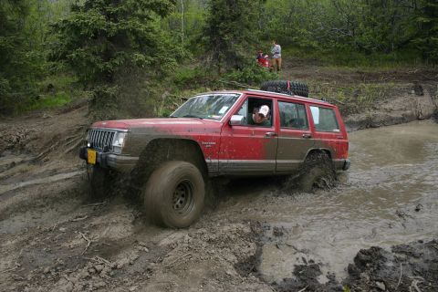 2003 Jeep TJ - A red Jeep Cherokee XJ navigates a muddy trail with a spare tire mounted on its roof.