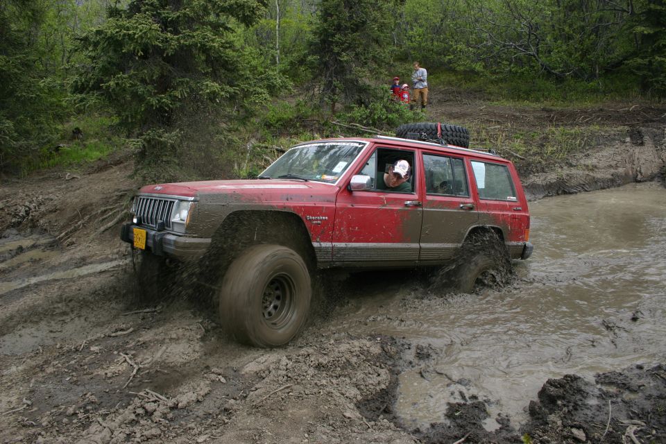 Red Jeep Cherokee XJ driving through mud, driver visible, spare tire on roof