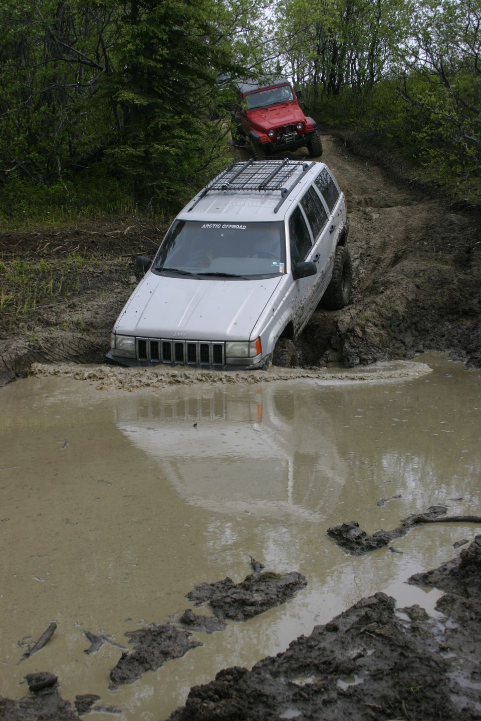 Silver Jeep Grand Cherokee stuck in mud puddle, red Jeep Wrangler behind it on a muddy trail