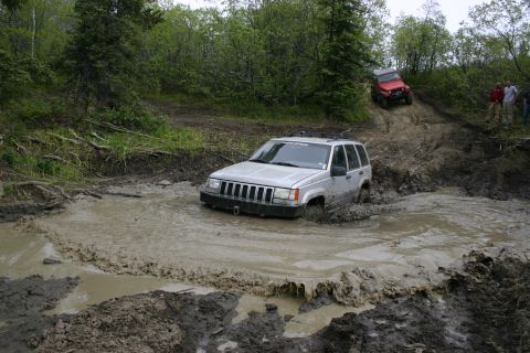 2003 Jeep TJ - A silver Jeep Grand Cherokee navigates a deep mud pit while a red Jeep Wrangler ascends a muddy incline in the background.