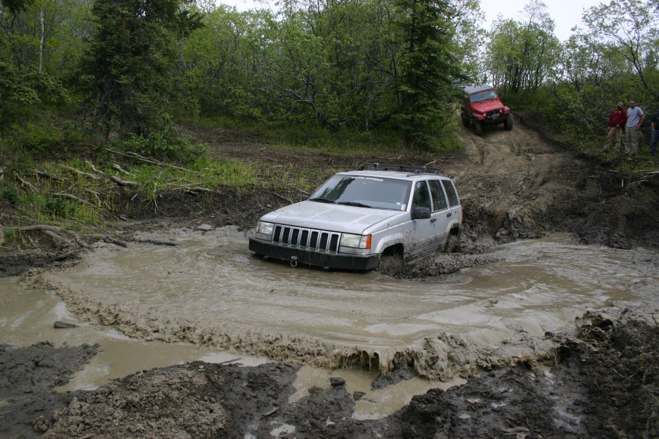 Silver Jeep Grand Cherokee stuck in mud puddle, red Jeep Wrangler on hill behind