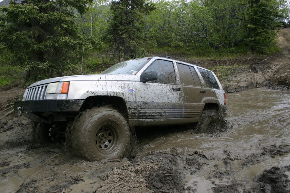 Silver SUV with large tires driving through mud, front grille visible