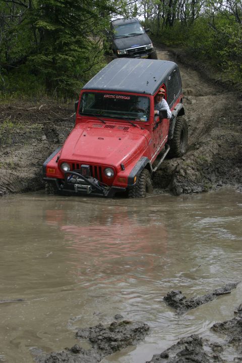 2003 Jeep TJ - A red Jeep Wrangler tackles a deep mud puddle on a wooded trail, followed by a dark SUV.