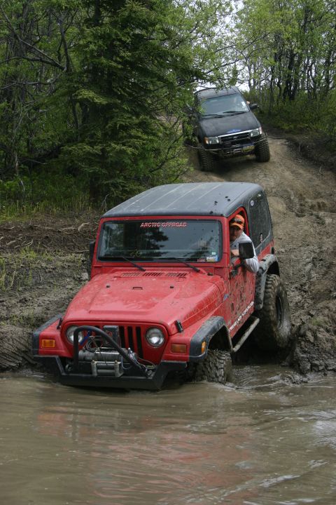 2003 Jeep TJ - Two off-road vehicles navigate a muddy trail, with a red Jeep TJ in the foreground and a black SUV following.