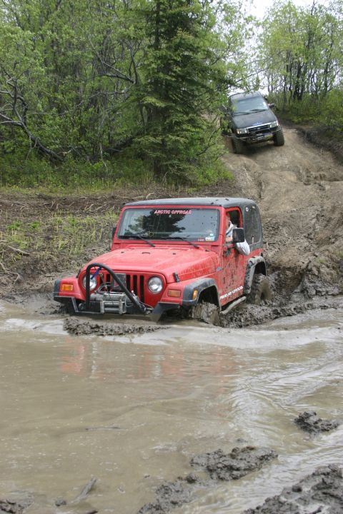 2003 Jeep TJ - Two off-road vehicles navigate a muddy trail with dense trees on either side.