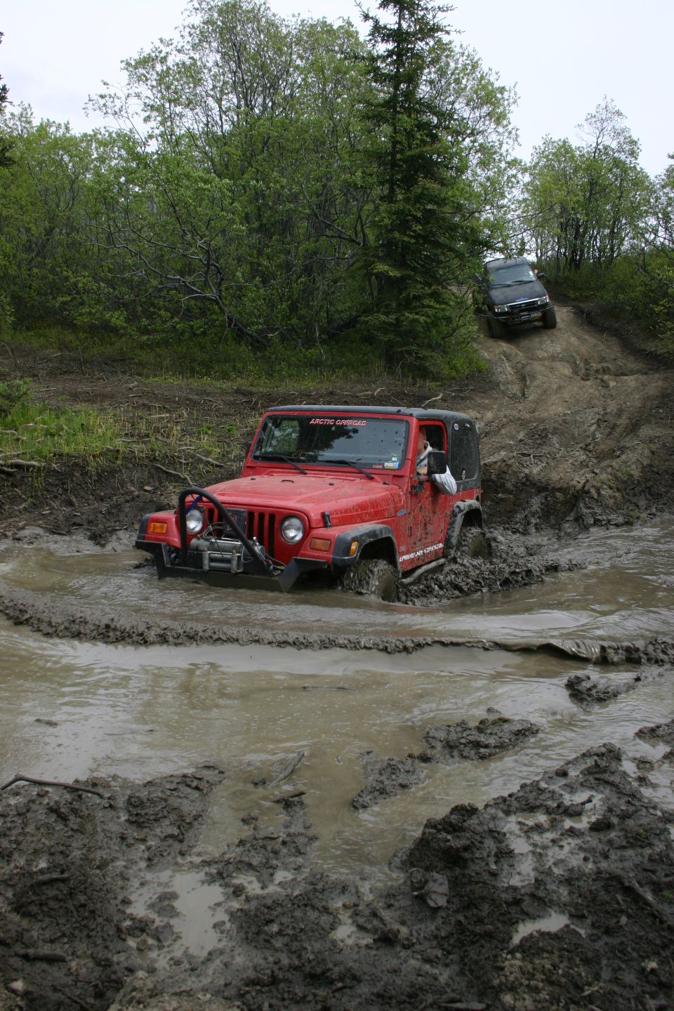 Red Jeep Wrangler stuck in mud, front bumper and winch visible. Another black SUV climbs a muddy hill in the background