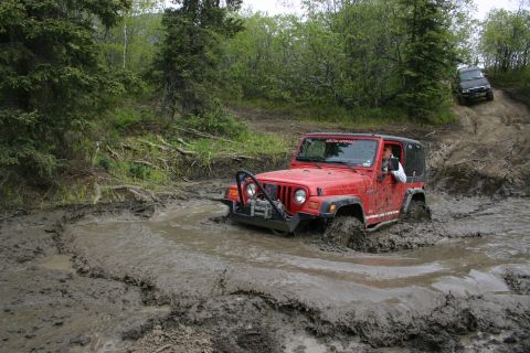 2003 Jeep TJ - A red 2003 Jeep TJ tackles a challenging mud obstacle while another vehicle ascends a muddy incline in the background.