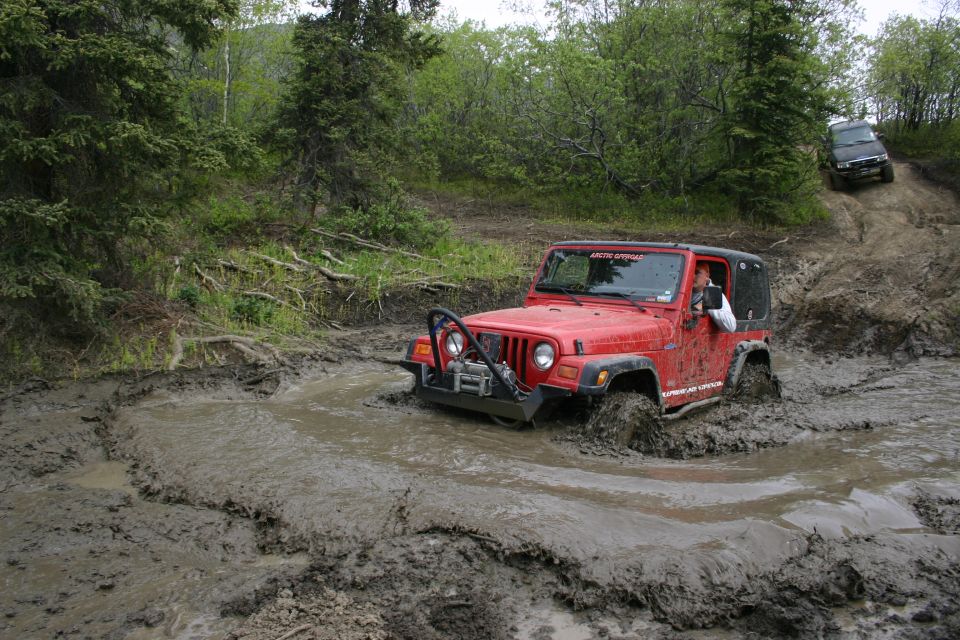 Red 2003 Jeep TJ navigating a deep mud pit with another SUV on a hill behind