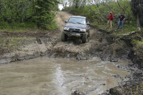 2003 Jeep TJ - A black pickup truck tackles a challenging muddy terrain with two onlookers observing the action.