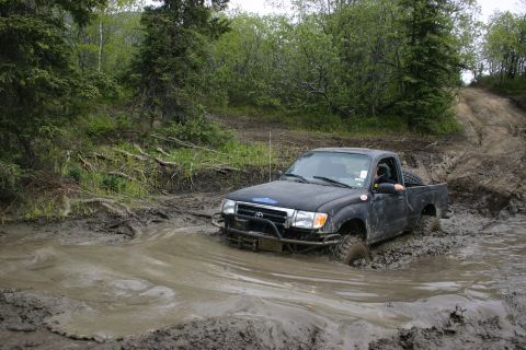 2003 Jeep TJ - A black Toyota Tacoma navigates a challenging muddy off-road trail, with trees and dense foliage surrounding the path.
