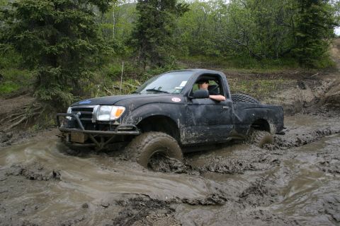 2003 Jeep TJ - A black pickup truck navigates a challenging muddy terrain, with its tires deep in the mire.