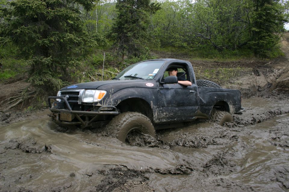 Black pickup truck stuck in mud, front bumper visible, driver in cab, trees and dirt track in background
