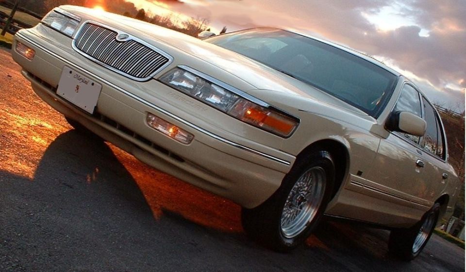 Cream Mercury Grand Marquis sedan, low front 3/4 view with wire wheels at sunset