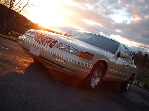 1996 Mercury Grand Marquis - A tan Mercury Grand Marquis is parked on a road as the sun sets behind it, casting a warm glow.