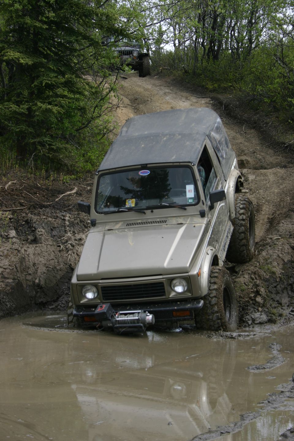Tan Suzuki Samurai with a soft top driving through mud, winch on front bumper, another vehicle visible on a hill behind