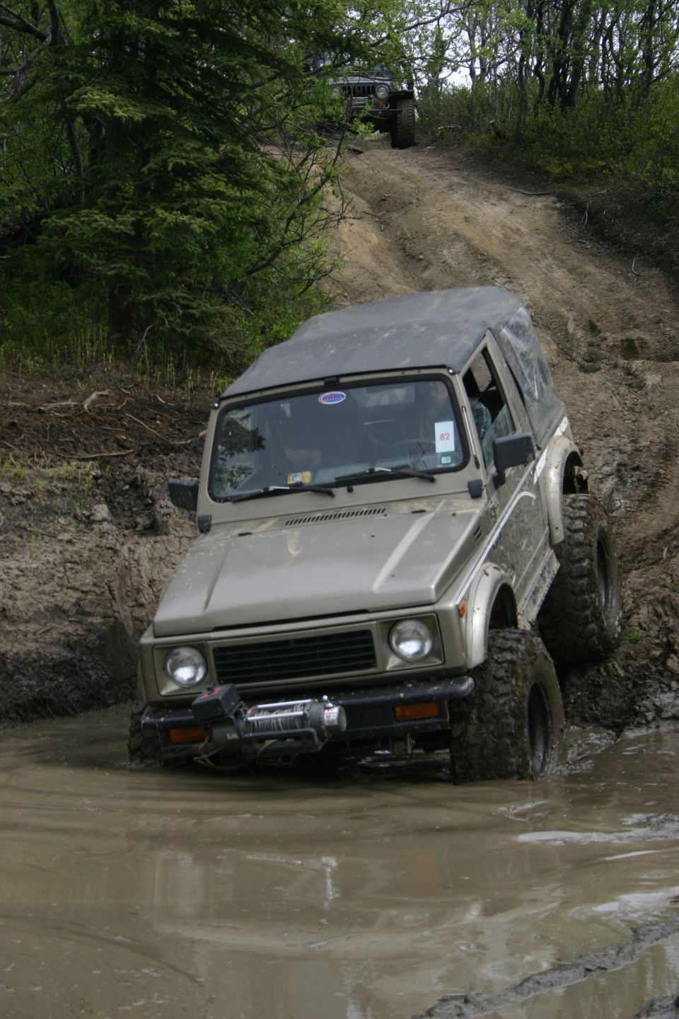 Tan off-road vehicle with soft top navigating a muddy trail, another vehicle visible uphill