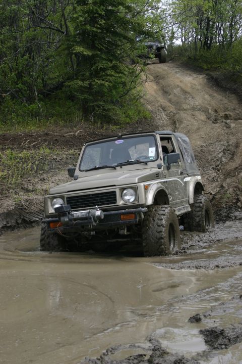 2003 Jeep TJ - Suzuki Samurai navigates a muddy off-road trail with another vehicle in the background.